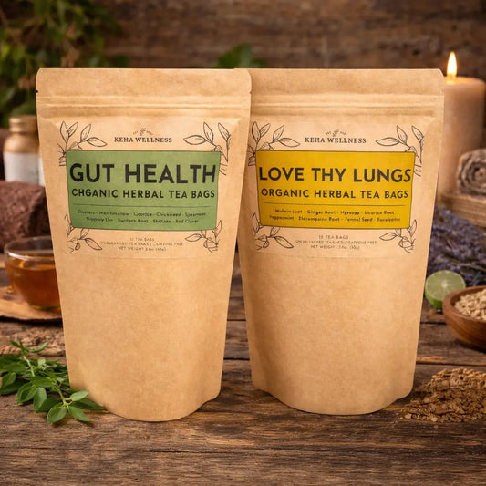 Two packages of organic herbal tea bags from Keha Wellness on a wooden surface with a candle and herbs in the background.